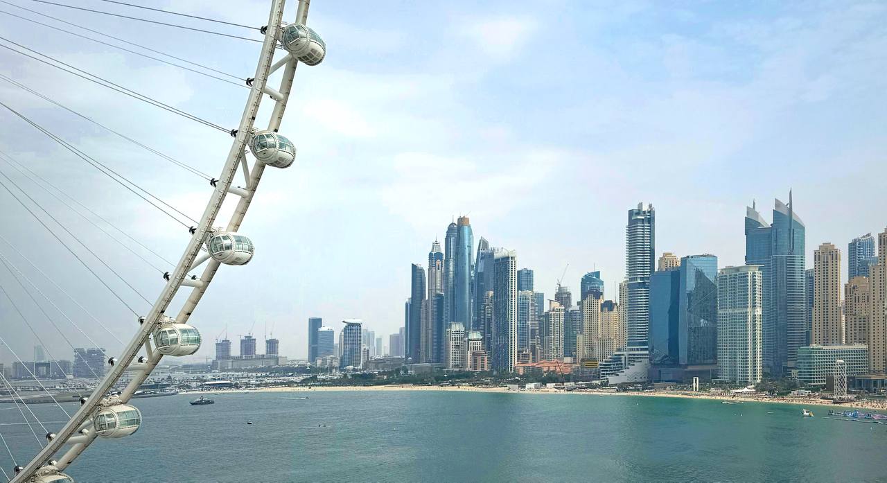 Part of a giant white observation wheel on the left, with a dense group of glass towers across green water.