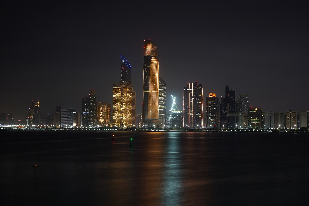 Lit towers along a waterfront at night, their lights reflected in still dark water.