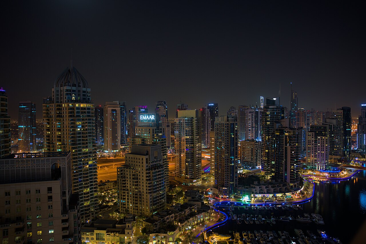 A dense group of lit skyscrapers at night around a curving waterway edged with blue lights.