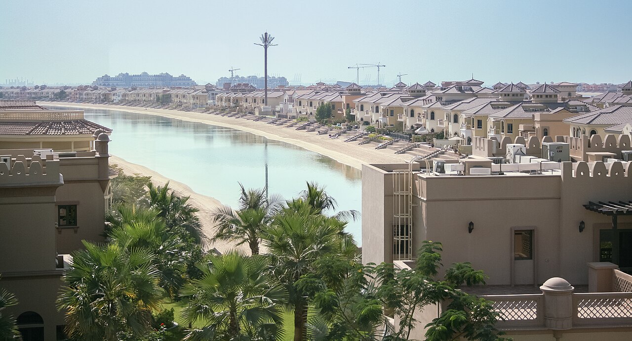 A curving row of sand-coloured villas facing a narrow beach and calm water, with palm trees in front.