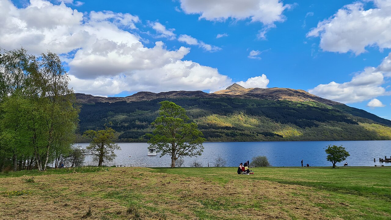 A grassy shore with a few trees beside a loch, with a green mountain and white clouds behind.