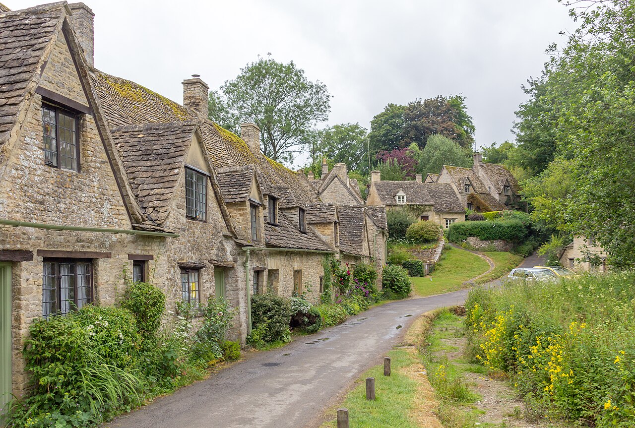 A lane curving past a terrace of old stone cottages with steep roofs, with wildflowers on the verge.