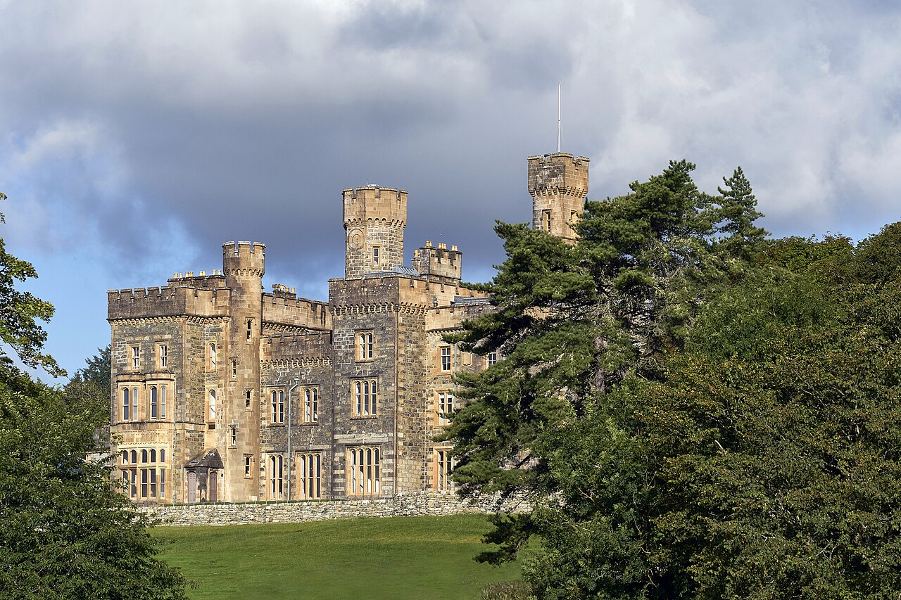 A large stone castle with round towers sitting on a grassy hill surrounded by green trees.