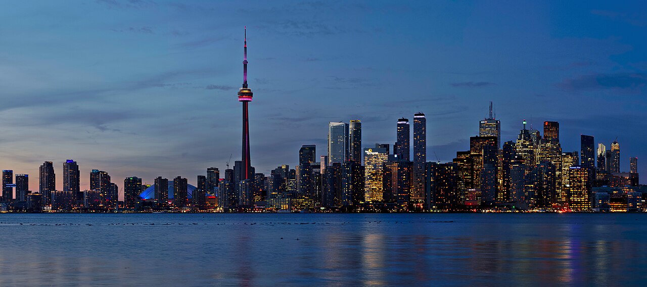 A city skyline with a tall slender tower lit in red, reflected in calm blue water at dusk.