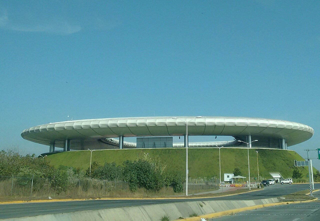 A low round stadium with a white ring-shaped roof set on a grass-covered mound under a clear sky.