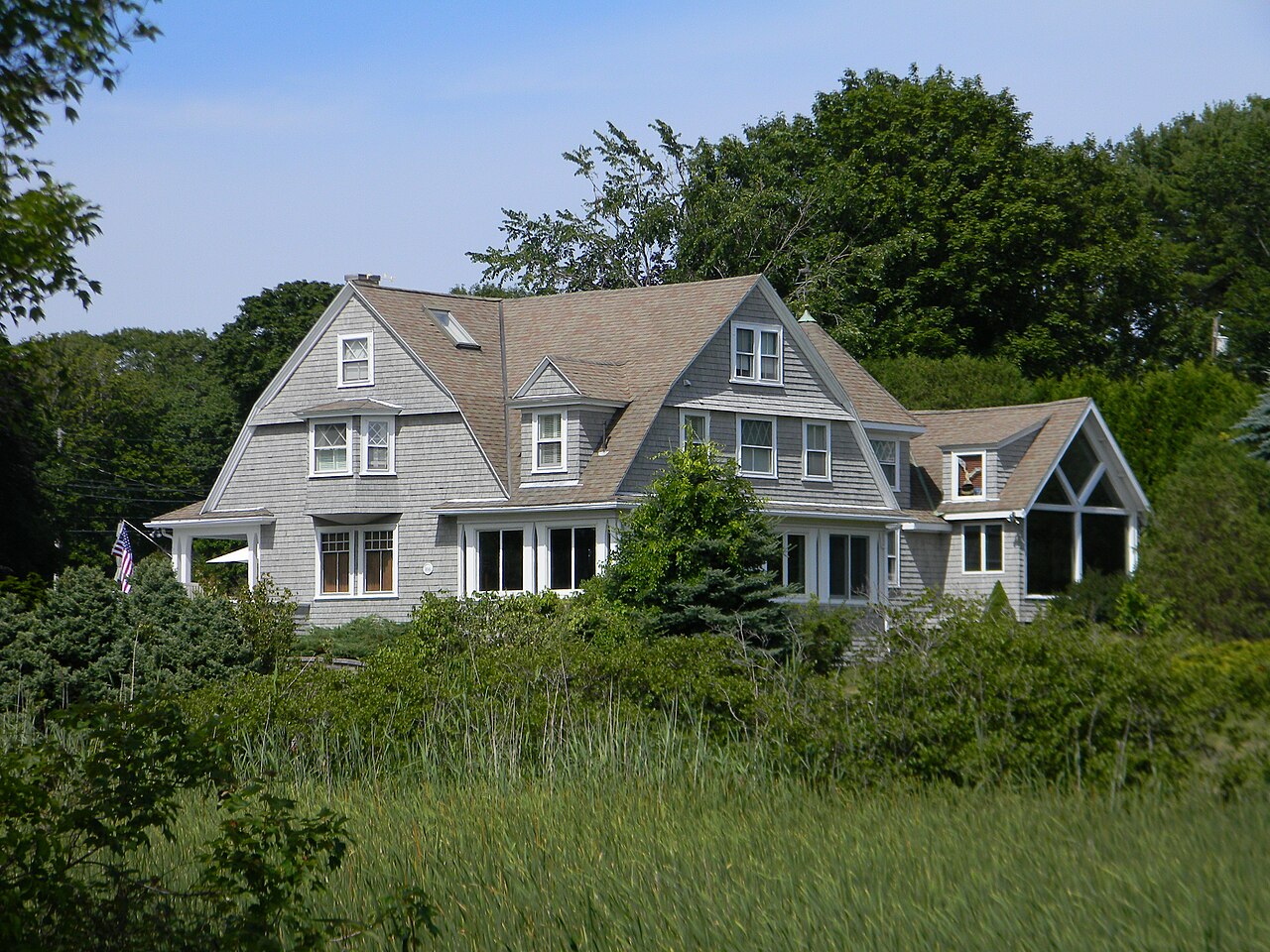 A large grey shingled house with several gables stands behind tall grass and shrubs.