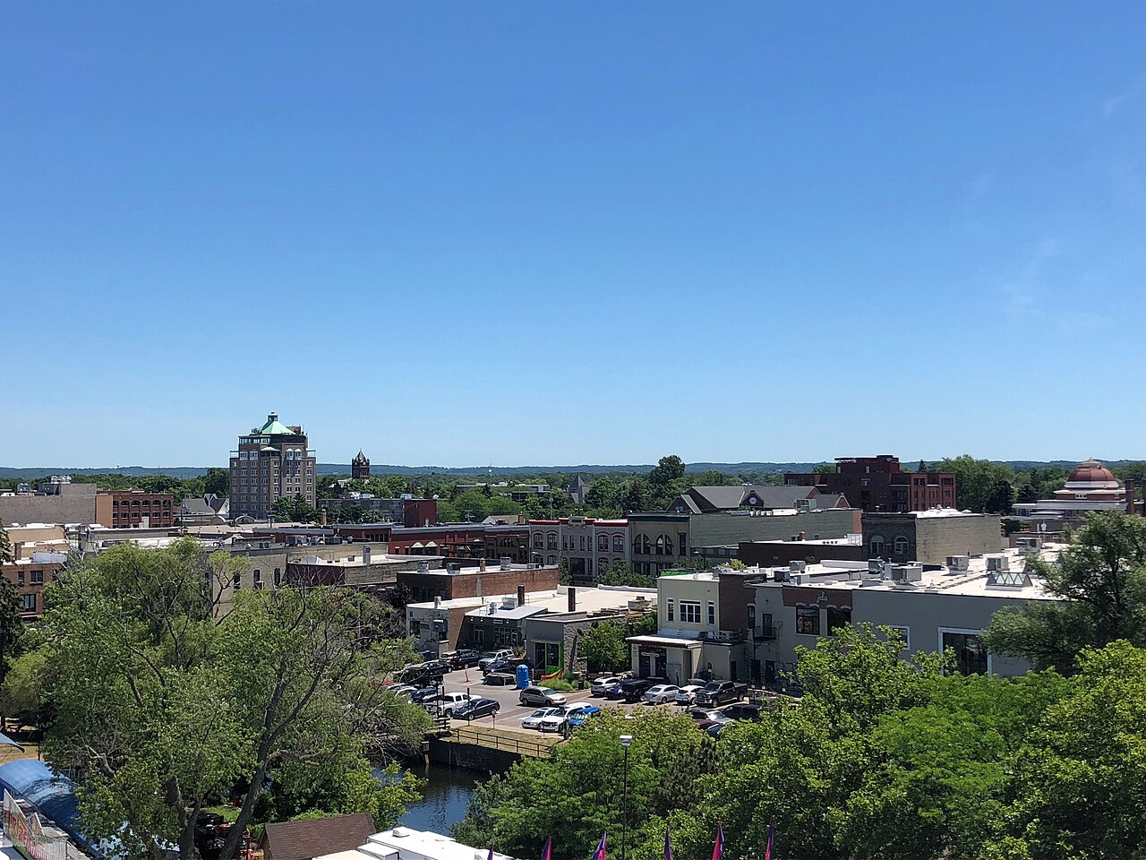 Low downtown buildings and a river behind a line of green treetops under a clear blue sky.
