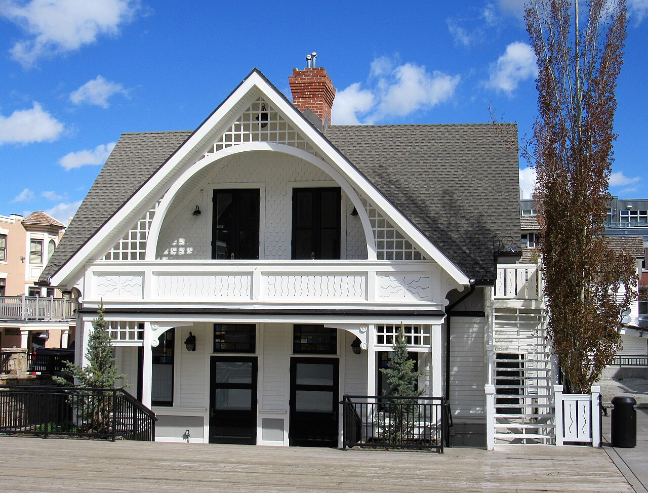 A white two-storey wooden building with a steep gabled roof, a balcony and black-trimmed doors.