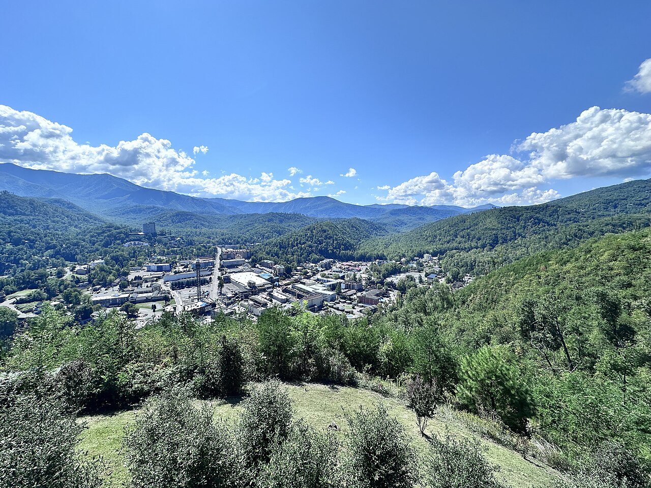 A small town fills a valley floor between forested ridges under a blue sky with white clouds.