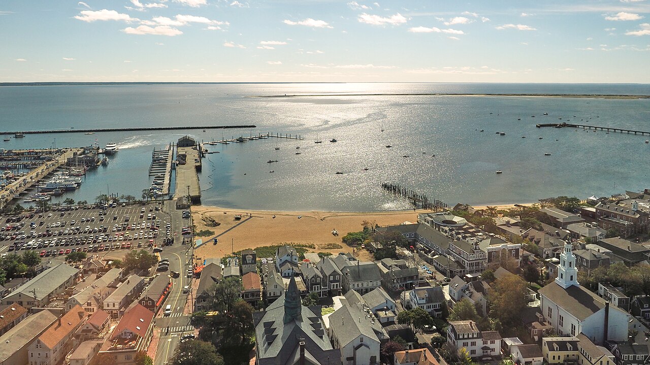Rooftops, a beach and long wharves reach into a wide sunlit harbour, seen from high above.