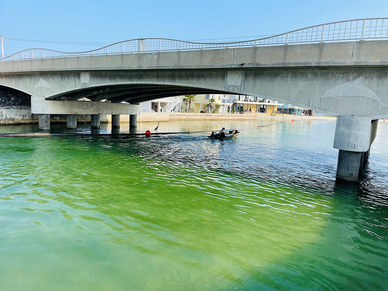 A low concrete road bridge crosses a channel of bright green water where a small boat passes.