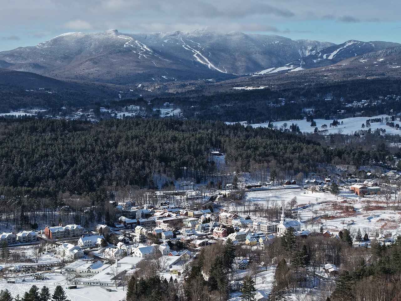 A snow-covered village lies among dark evergreen forest with ski runs on the mountains behind.