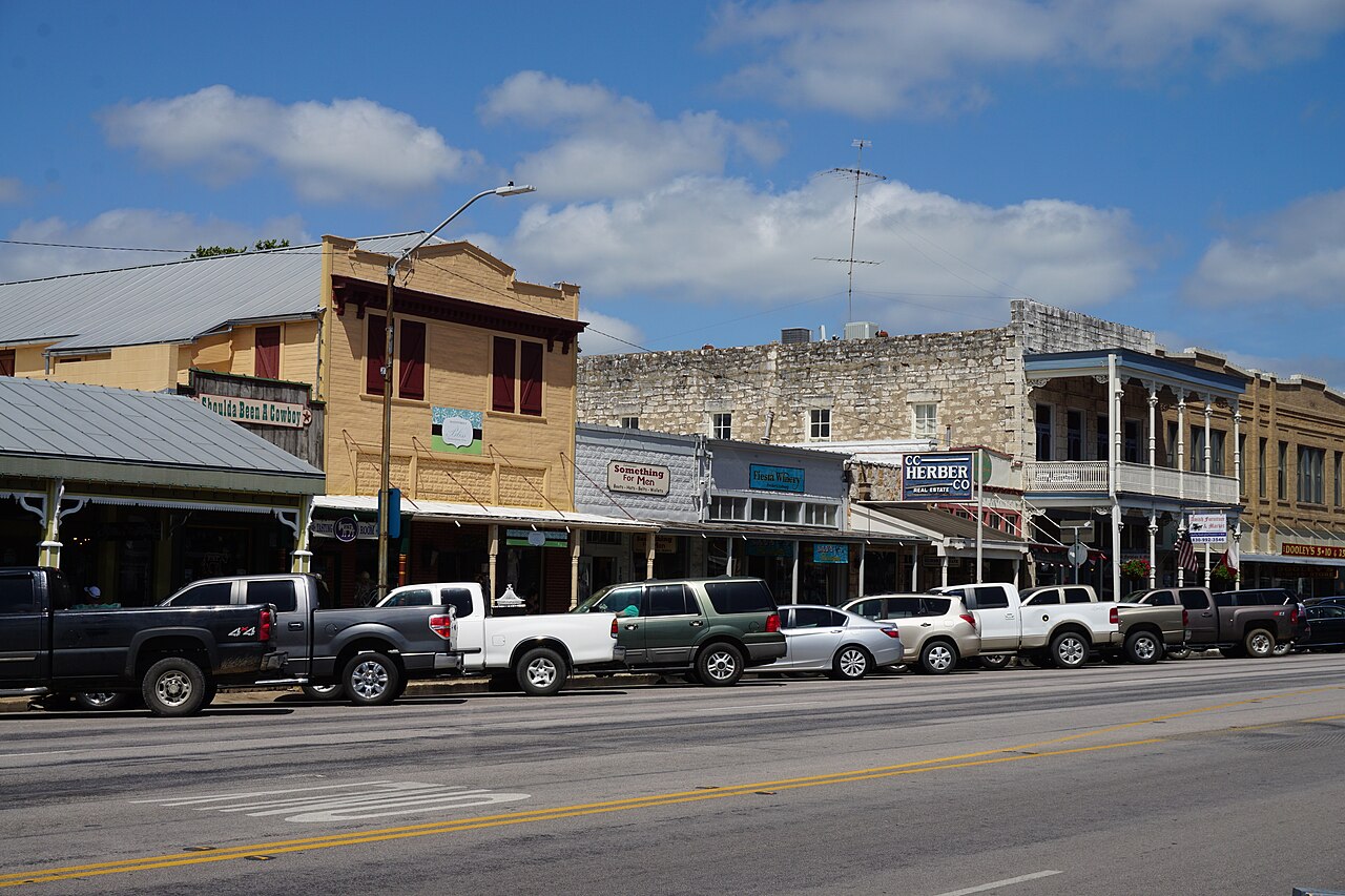 Old stone and timber shopfronts with covered sidewalks line a wide street with parked pickup trucks.