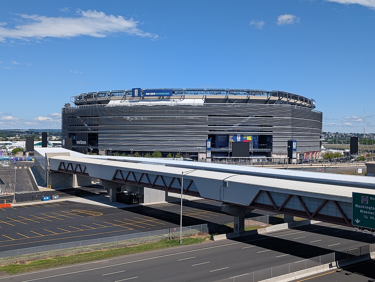 A large grey oval stadium behind an elevated walkway and empty roads under a clear blue sky.
