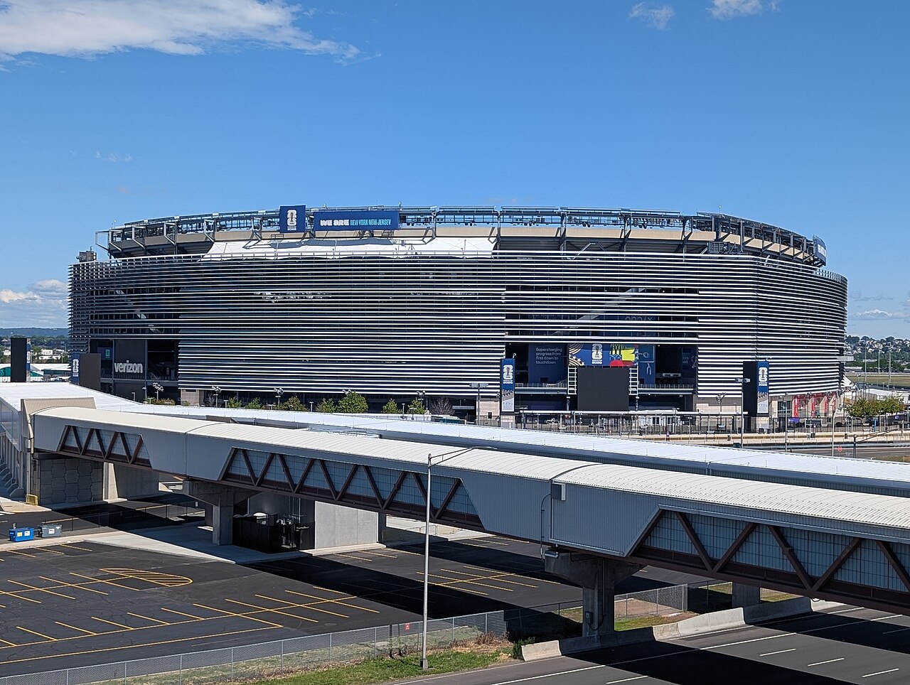 A grey oval stadium fills the frame above an elevated walkway, under a clear blue sky.