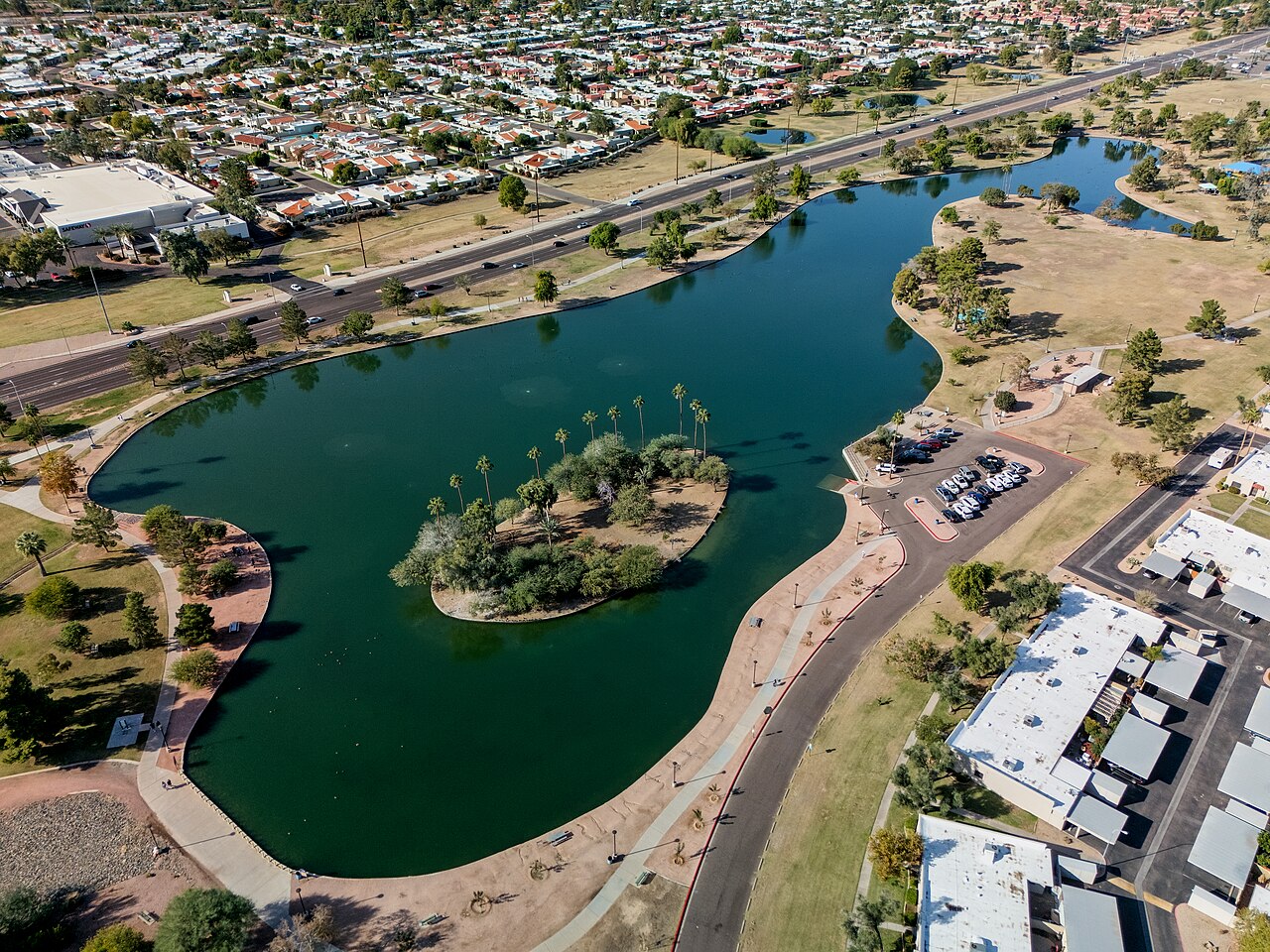 A dark green lake with a small tree-covered island, ringed by paths, lawns and low houses.