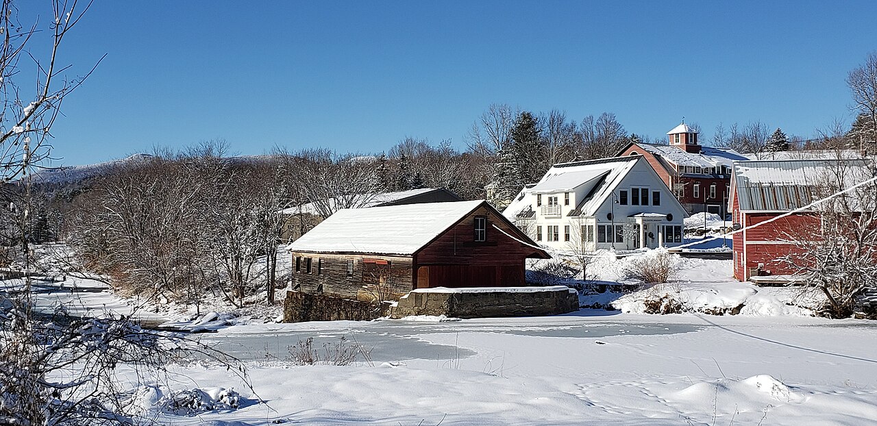 A red wooden mill and a white barn with a cupola stand beside a frozen river in deep snow.