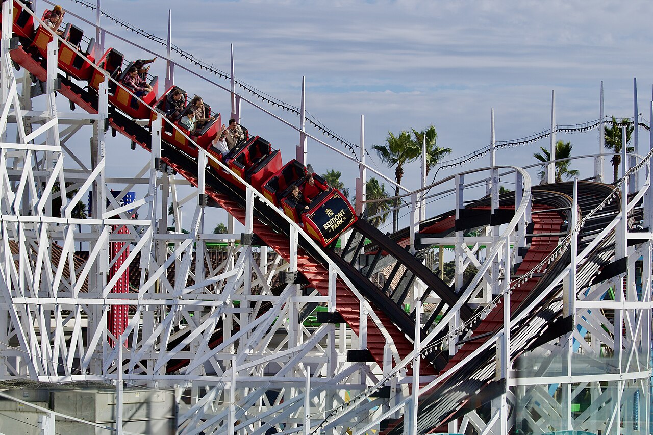 A red roller coaster train climbs a curve of white wooden lattice track with palm trees behind.