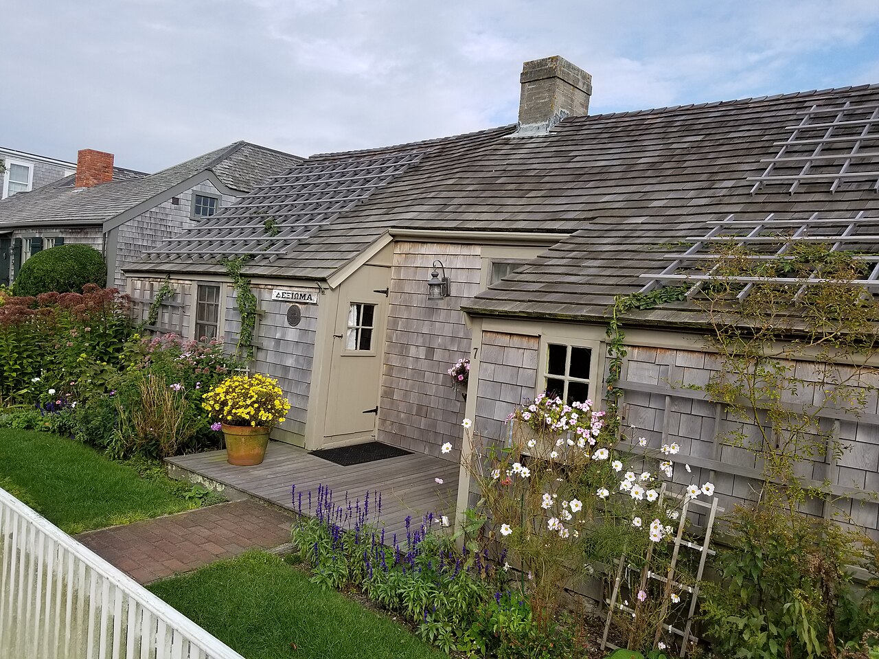 A low grey shingled cottage with a climbing trellis on its roof, behind a garden of white and yellow flowers.