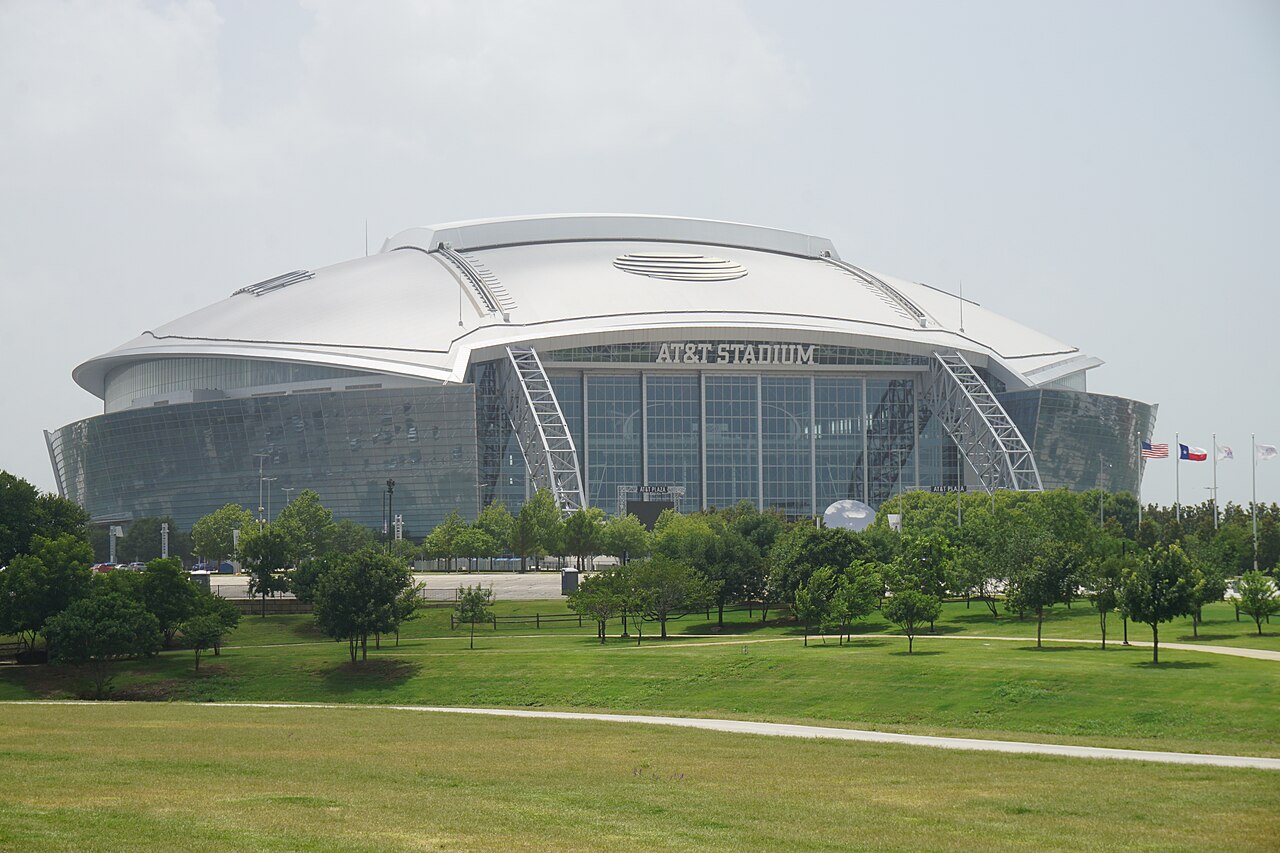 A large domed stadium with a white roof and glass end wall stands beyond a lawn dotted with young trees.