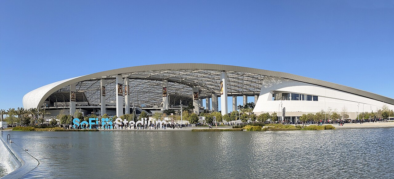 A sweeping curved stadium canopy on white columns rises behind a lake, with the stadium's name in large letters at the water's edge.