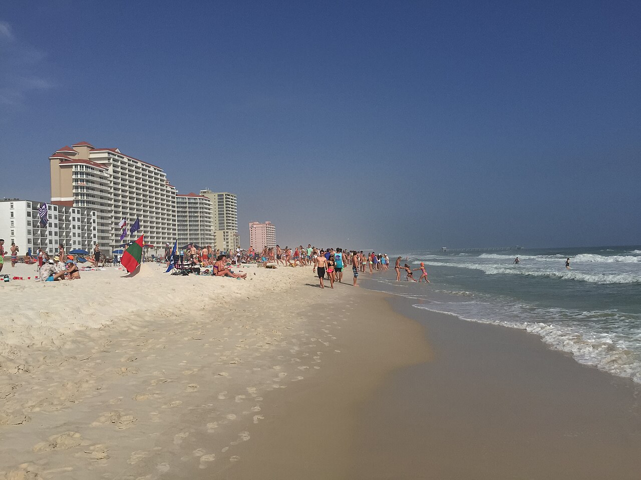 A crowded sandy beach curves away along the surf, with a row of tall pale apartment towers behind it in the haze.