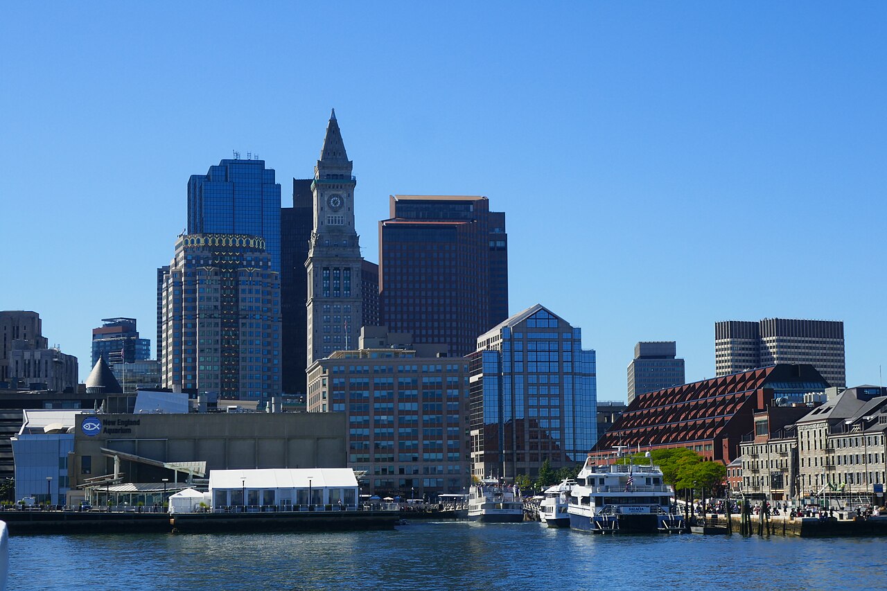 Glass office towers and an older clock tower stand behind a waterfront of wharf buildings and moored boats under a deep blue sky.