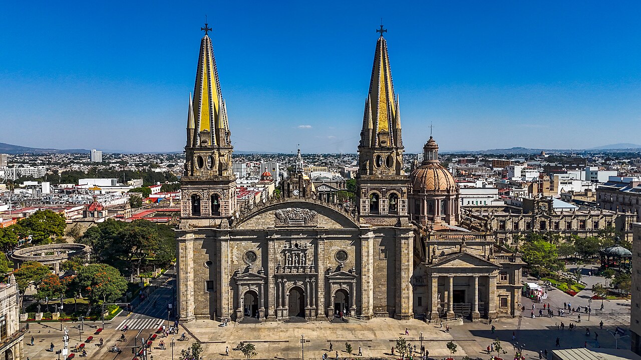 Two tall yellow-tiled spires rise from a stone cathedral above a plaza, with the low city spreading to the horizon under a blue sky.