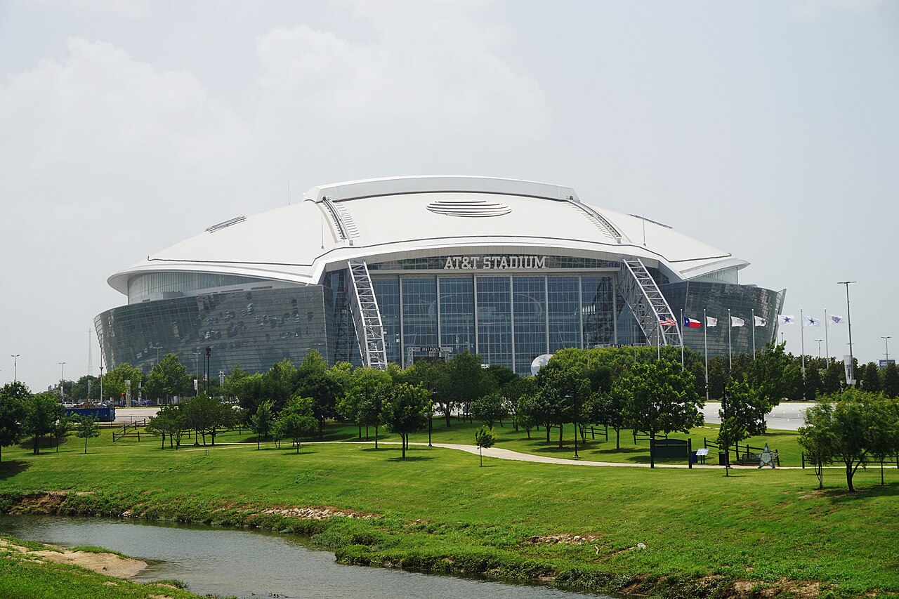 A white-roofed domed stadium with a glass end wall stands behind a green park with young trees and a small stream.