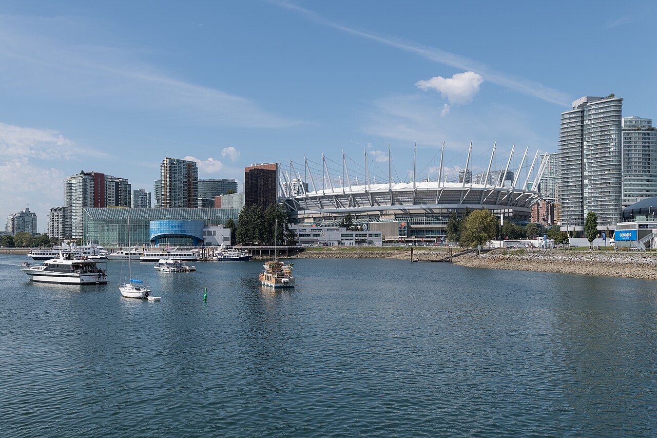 A low round stadium ringed with white roof masts sits on the far shore of a calm inlet, with boats at anchor and apartment towers on either side.